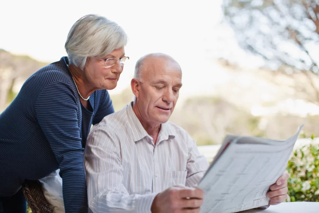 Older man reading a newspaper while an older woman reads over his shoulder